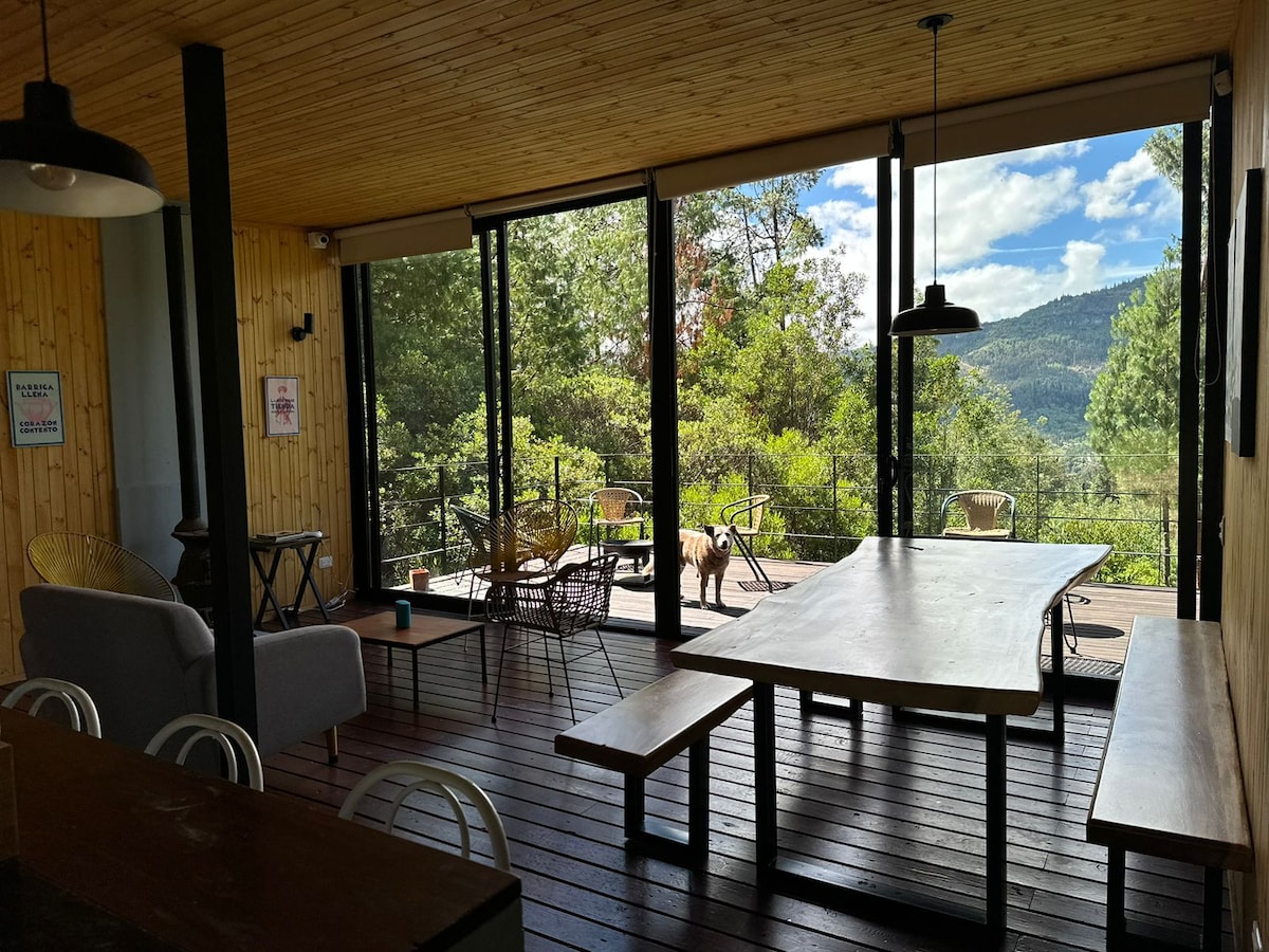 A spacious living area features large glass walls that open to a deck surrounded by greenery and mountains. Natural light illuminates the room, where a wooden dining table and benches sit center stage, complemented by a cozy seating area with a couch.