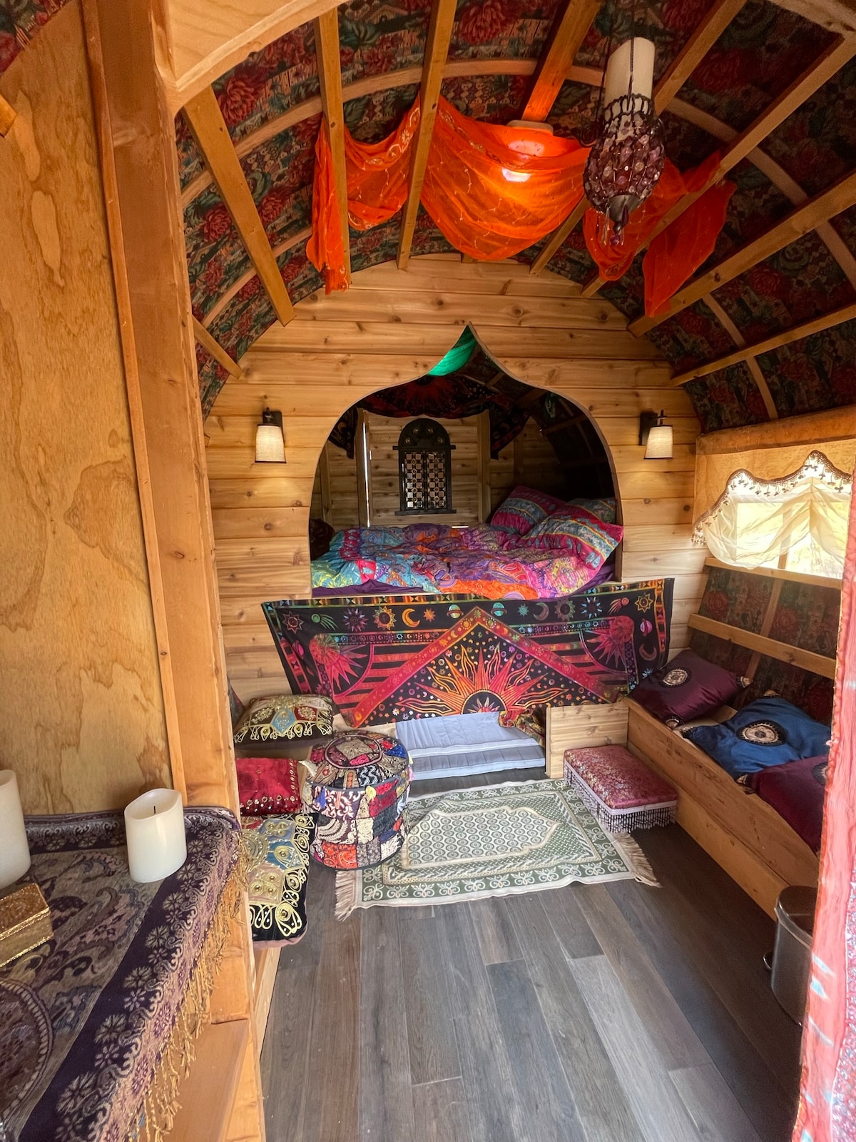 The interior of the caravan features wooden walls and an arched window above a queen-sized bed adorned with vibrant bedding. Colorful textiles and cushions create a cozy atmosphere, while soft lighting from the wall sconces enhances the charm of the space.