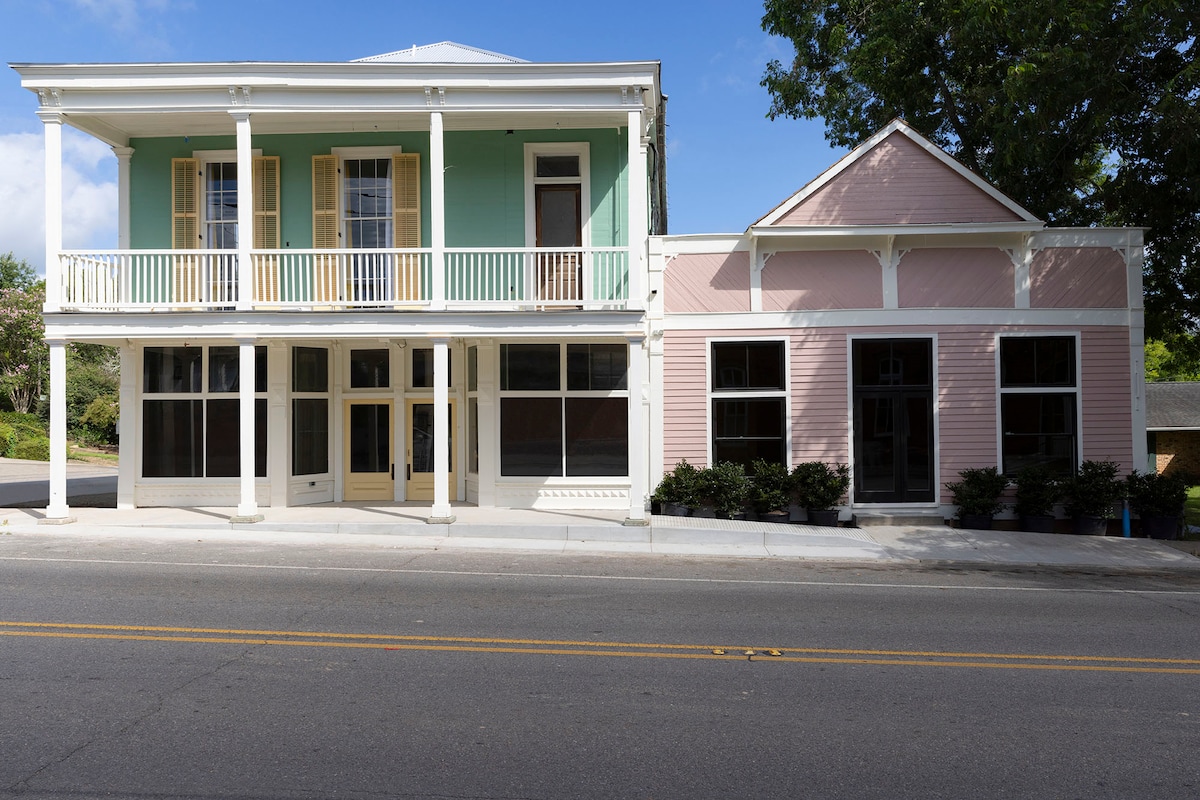 The exterior of Hotel Klaus presents a charming two-story building with a pastel green and pink facade. A welcoming porch with white railings is featured on the upper level, while large windows allow for natural light. The surrounding area is landscaped with tidy greenery.