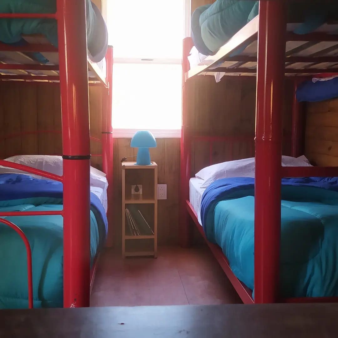 A shared sleeping area is furnished with two sets of metal bunk beds featuring red frames. Each bed is made with soft blue blankets, and a small wooden nightstand is placed between them, holding a blue lamp and some books. Natural light enters through a window.