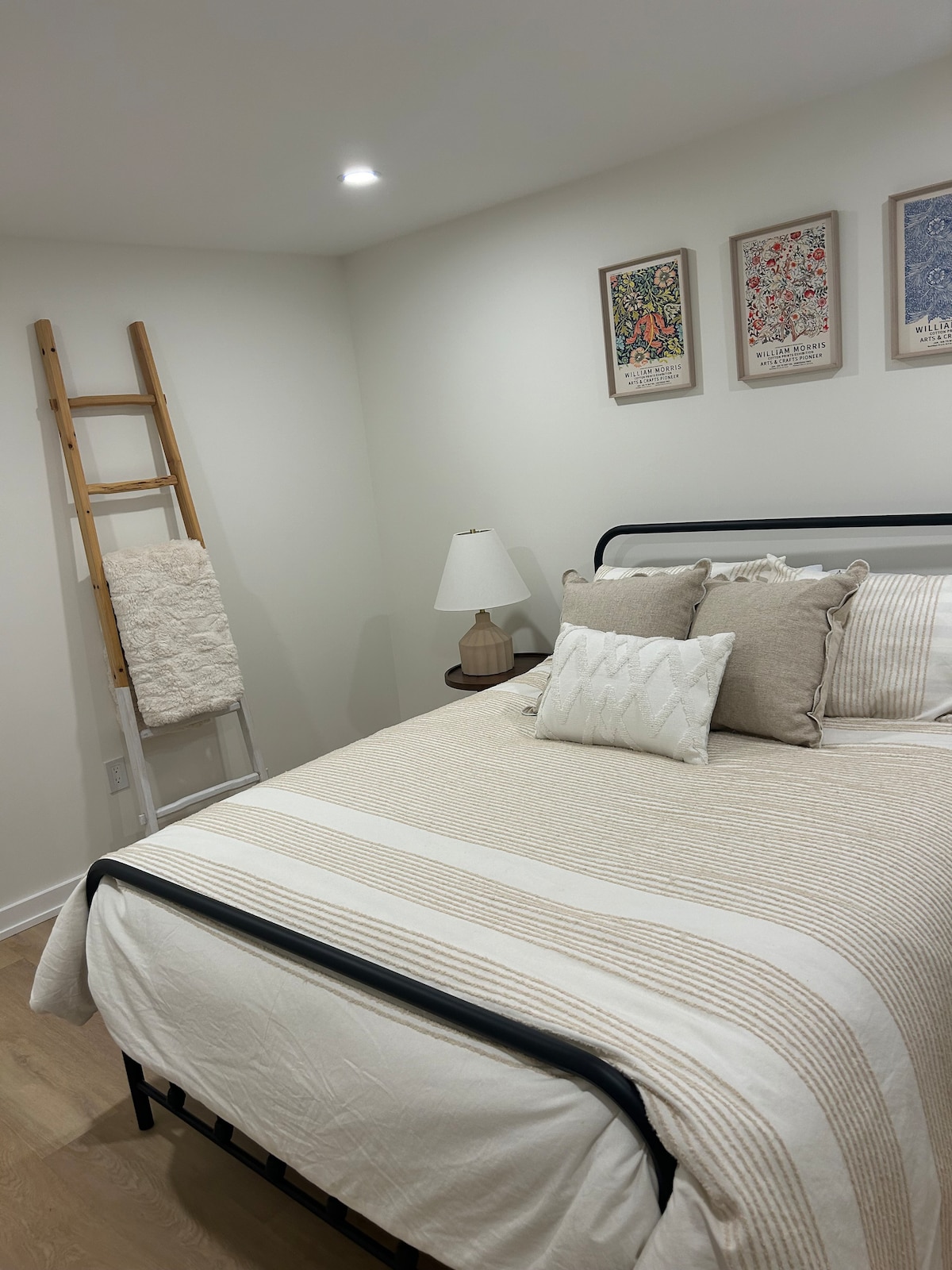 A serene bedroom features a spacious bed adorned with neutral-toned linens and decorative pillows. A soft light from a bedside lamp illuminates the area. Nearby, a wooden ladder displays a cozy throw, and vibrant artwork adorns the walls.