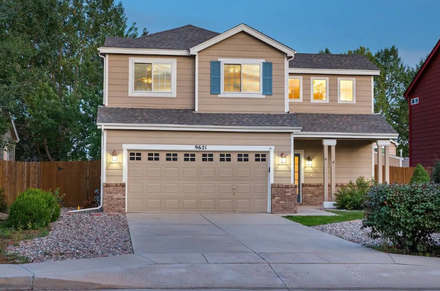The exterior of the home features a two-story design with a light brown facade and blue shutters. The driveway leads to a two-car garage, with stone accents and well-maintained landscaping, welcoming guests in a tranquil, residential setting.