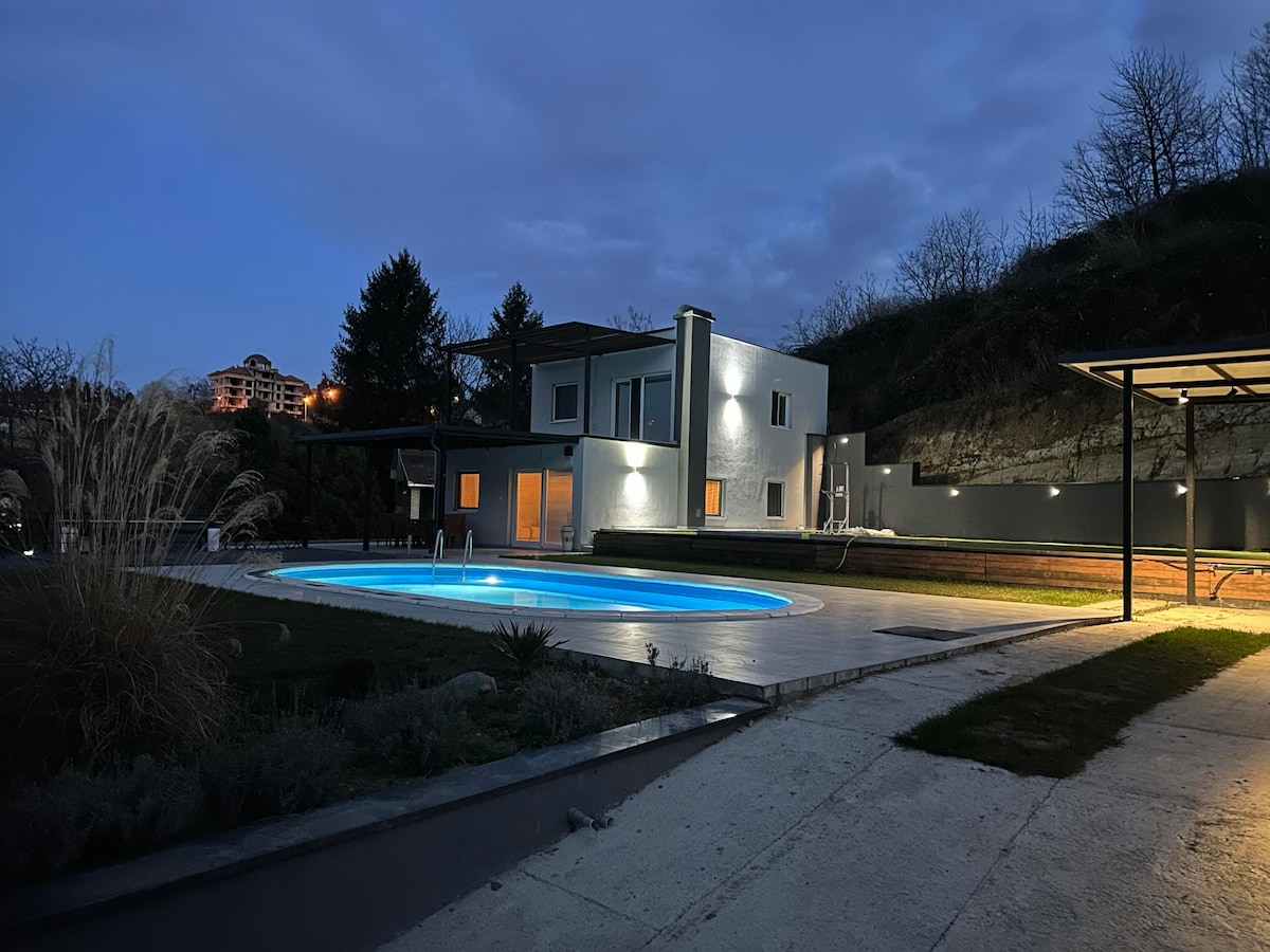 A modern building is shown at dusk, featuring large windows that reflect soft outdoor lighting. A circular swimming pool is positioned in the foreground, surrounded by manicured grass and decorative plants. The scenic hillside adds a tranquil backdrop, enhancing the peaceful outdoor space.