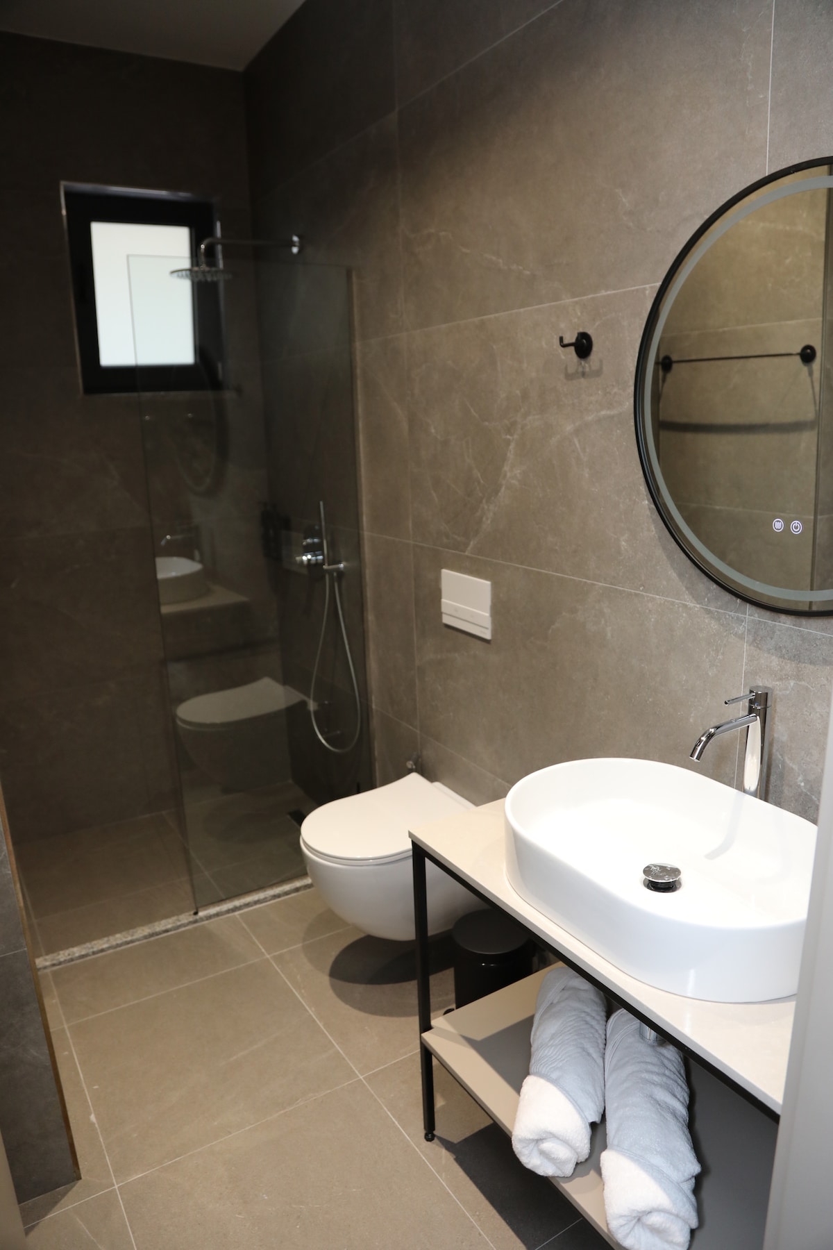 A modern bathroom features sleek gray tile walls and flooring. A walk-in shower with glass panels is complemented by a round mirror above a white sink. A shelf holds neatly rolled towels, adding to the clean and organized appearance.