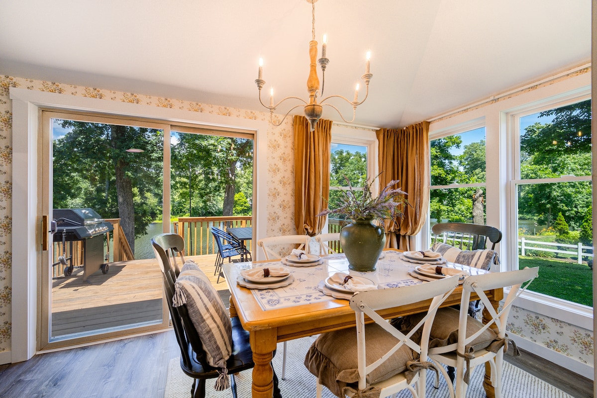 The dining area features a wooden table set for six, accompanied by white chairs. Natural light floods through large windows, enhancing the view of the outdoor deck and surrounding greenery. A vase of flowers adds a touch of elegance, while warm curtains frame the scene.