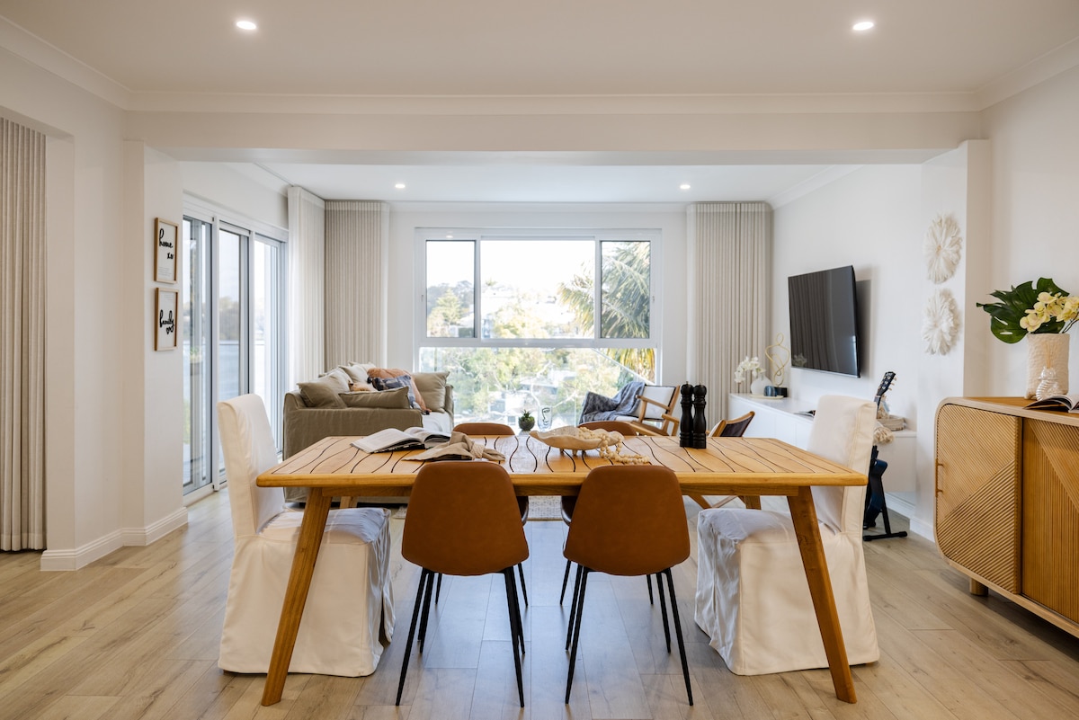 A modern dining area features a long wooden table surrounded by brown chairs with white covers. Large windows provide ample natural light and offer views of the greenery outside. A comfortable living area is visible in the background, with soft furnishings and a wall-mounted television.