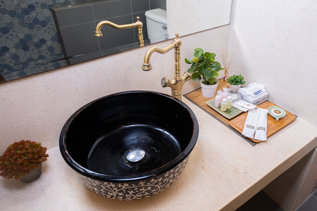 A round black sink with intricate designs is positioned on a light-colored countertop. A stylish brass faucet complements the sink. A small potted plant and a wooden tray with toiletries are neatly arranged beside it, creating a well-organized and functional space.