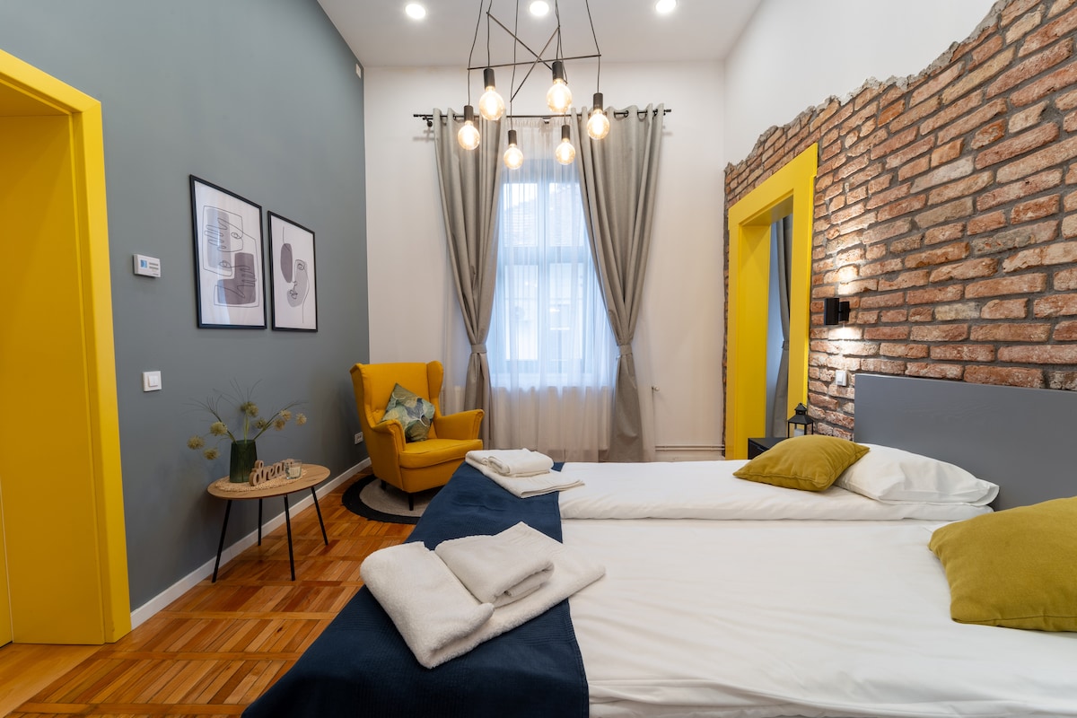 A modern studio features exposed brick accents and a striking design. Two spacious beds are arranged with rolled linens and decorative pillows. A yellow armchair complements the vibrant decor. Large windows allow natural light to illuminate the room, enhanced by stylish light fixtures.