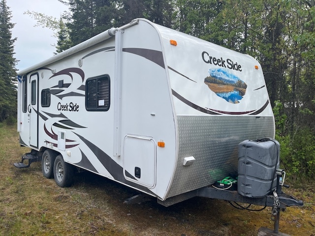 A camper is parked outdoors, featuring a streamlined exterior design with a compact kitchen area and a large window. The neutral color palette consists of white with brown and gray accents. The surrounding landscape includes grassy terrain and trees, setting a natural backdrop.