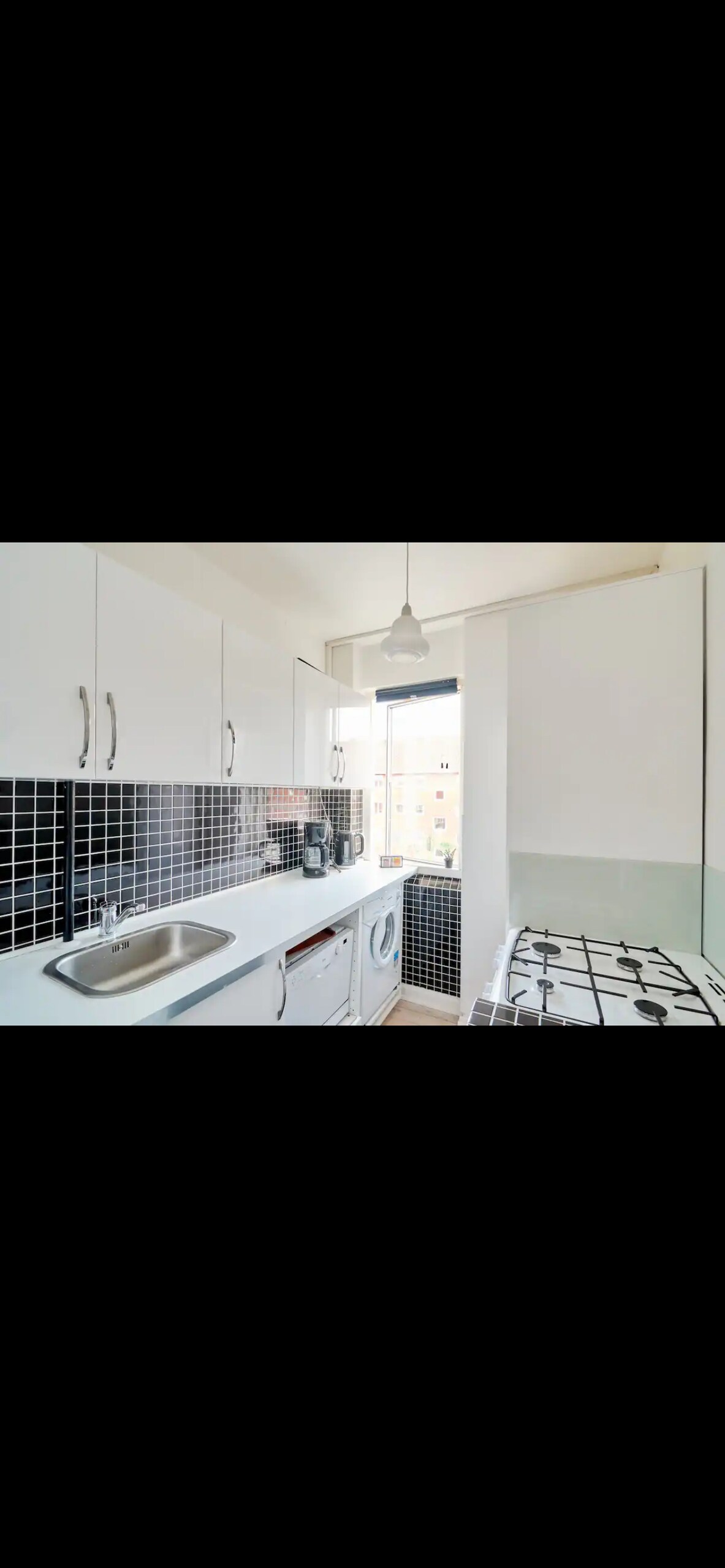 A bright kitchen area is equipped with modern appliances, including a washing machine and a gas stove. The cabinets are white, complemented by a black and white tiled backsplash. Natural light enters through a window, creating an airy atmosphere.