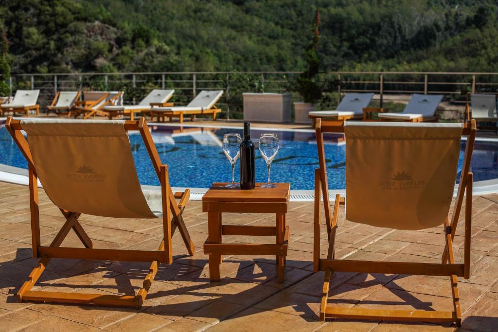 Two wooden lounge chairs are positioned beside a swimming pool, with a small table between them. Two glasses and a bottle are placed on the table. The pool is surrounded by greenery, creating a relaxing outdoor environment.