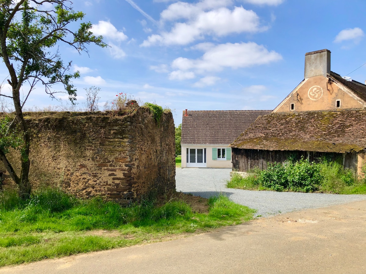 The image captures a charming view of the property, showcasing a rustic stone wall on the left and a traditional farmhouse with a slate roof on the right. The gravel driveway leads to the house, surrounded by lush greenery and a clear blue sky.