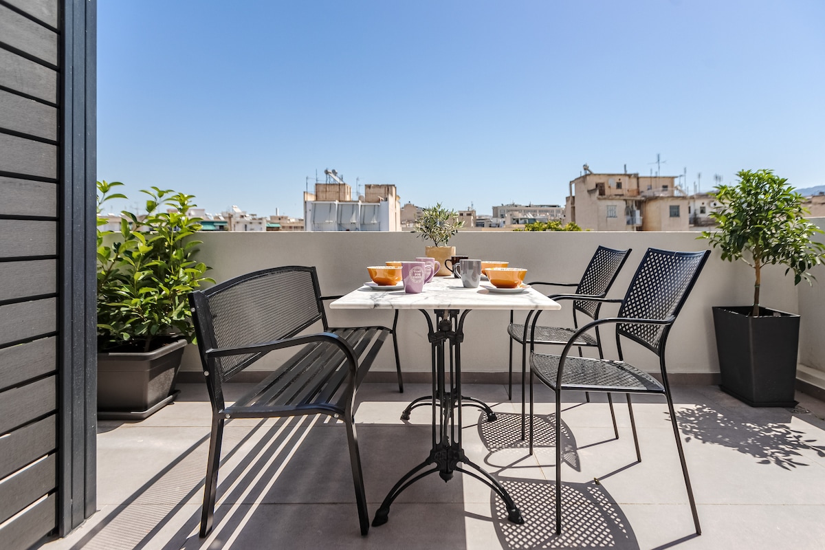 An outdoor terrace features a table set for four with vibrant bowls and cups. Black metal chairs surround the table, and potted plants are placed in each corner, enhancing the view of the Athenian skyline under clear blue skies.