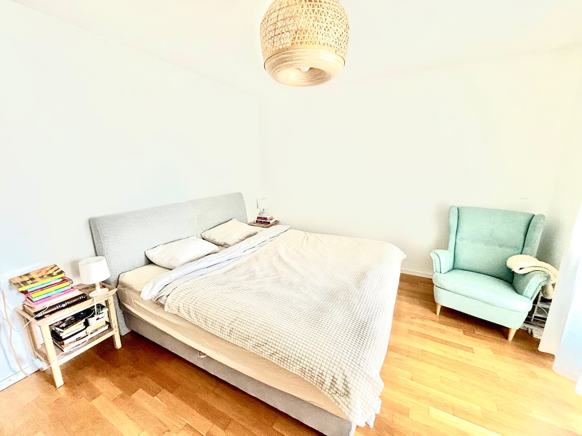 A light-filled bedroom features a spacious bed with a neutral-colored blanket, accompanied by a bedside table with books. An accent chair in a soft blue hue adds a cozy touch. The room is characterized by wooden flooring and soft white walls.