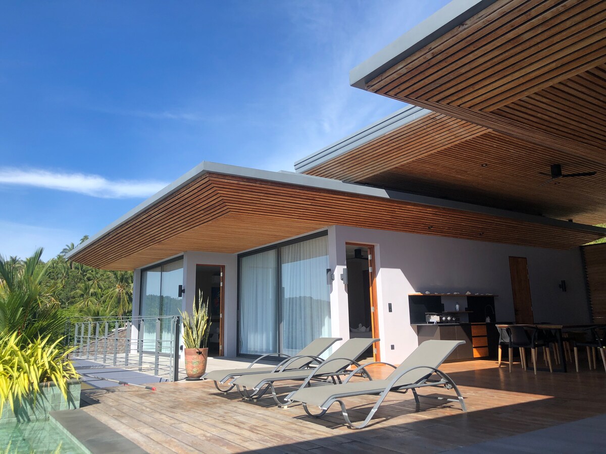 A modern villa exterior is presented, featuring a spacious wooden deck with sleek lounge chairs aligned for relaxation. Large glass doors provide access to the indoor living space, flanked by tropical greenery. A shaded overhang with wooden paneling completes the inviting design.