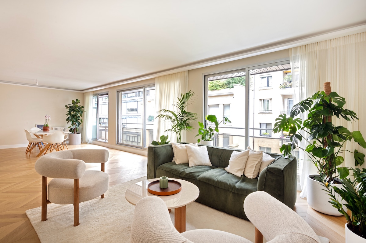 A spacious living area features large windows allowing ample natural light. A plush green sofa is surrounded by potted plants, while a round coffee table sits at the center. Dining space with a table and chairs can be seen in the background.