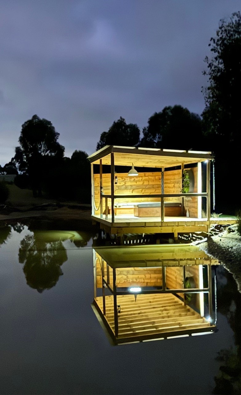 A wooden structure is illuminated at night, sitting on the edge of a calm body of water. The building's reflection creates a serene symmetry, showcasing its exterior features and ambient lighting amidst the surrounding trees.
