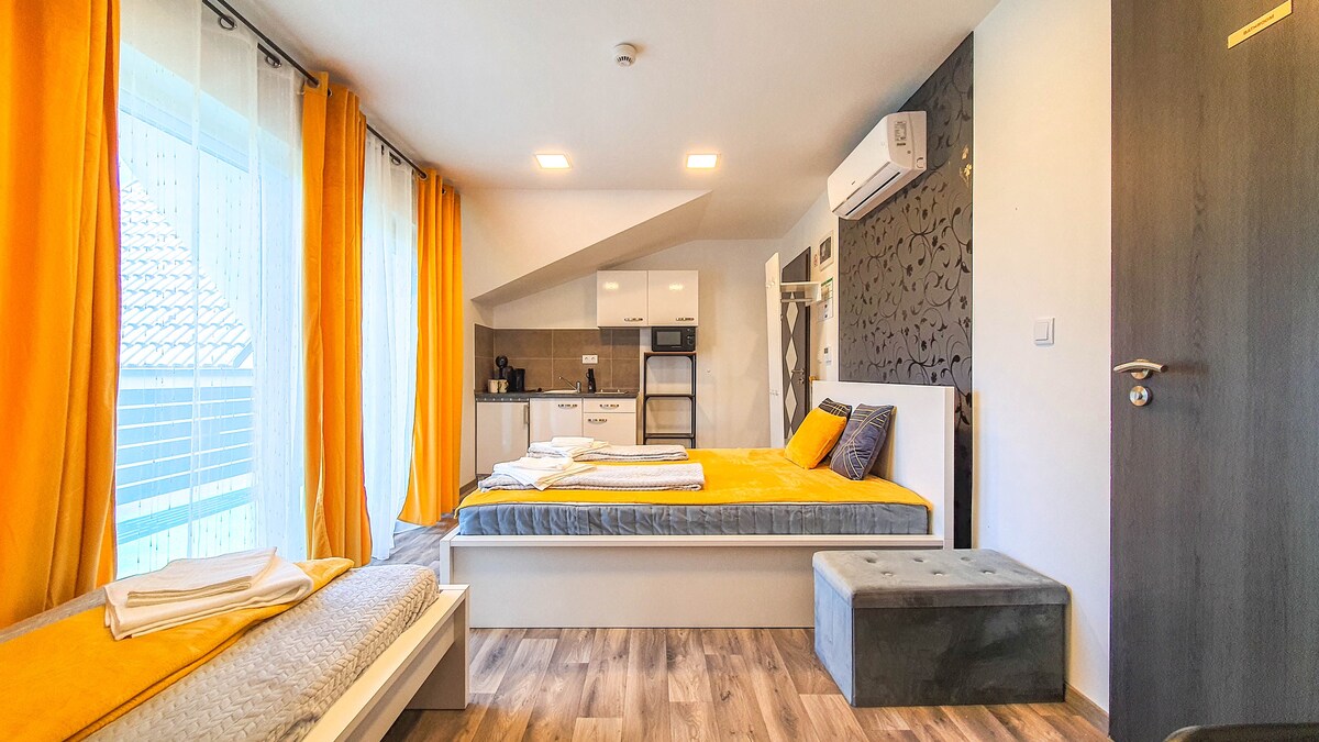 A bright room is presented, featuring a spacious bed dressed in vibrant yellow and gray linens. Large windows with sheer curtains allow natural light to fill the space. A modern kitchenette with appliances is visible, along with a comfortable seating area.