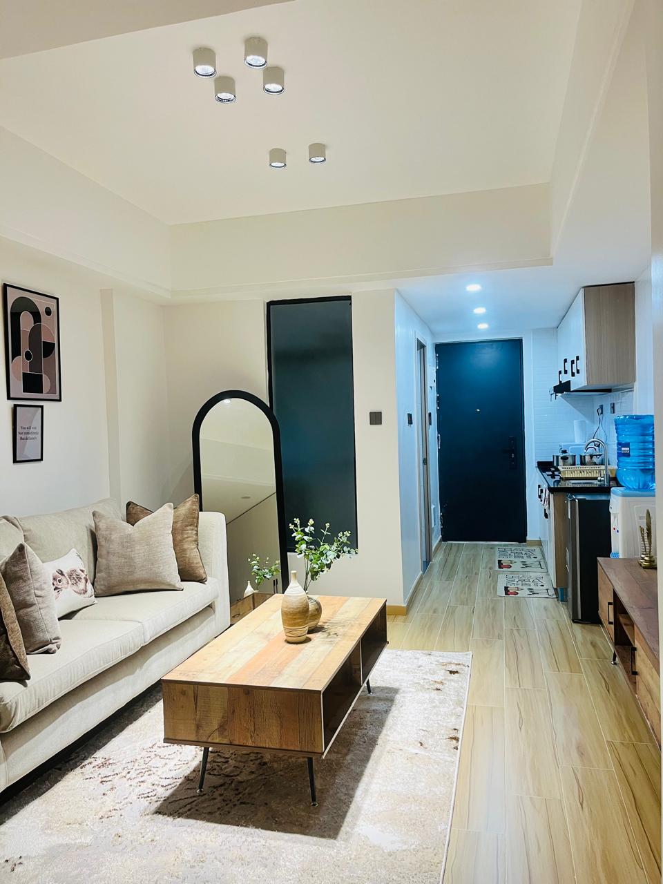 A cozy living area features a light-colored sofa adorned with decorative cushions. A wooden coffee table sits on a soft area rug, with a simple vase of greenery displayed on top. Warm lighting is provided by recessed ceiling fixtures, enhancing the space.