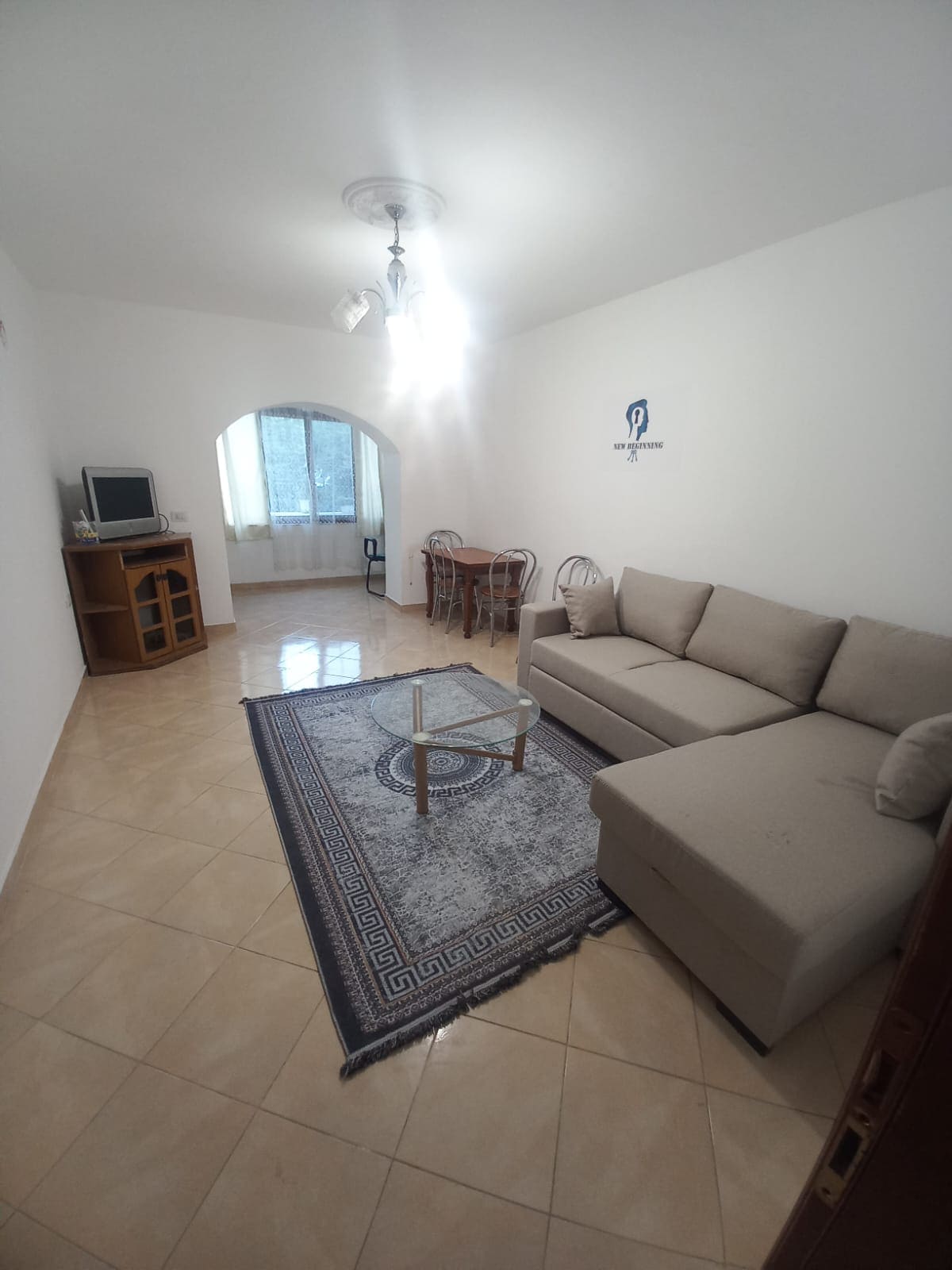 A light-filled living area is characterized by a sectional sofa in a neutral shade. A small wooden table is accompanied by four dining chairs. A patterned rug adds texture to the tile floor, while a television unit occupies one corner. Natural light enters through an arched window.