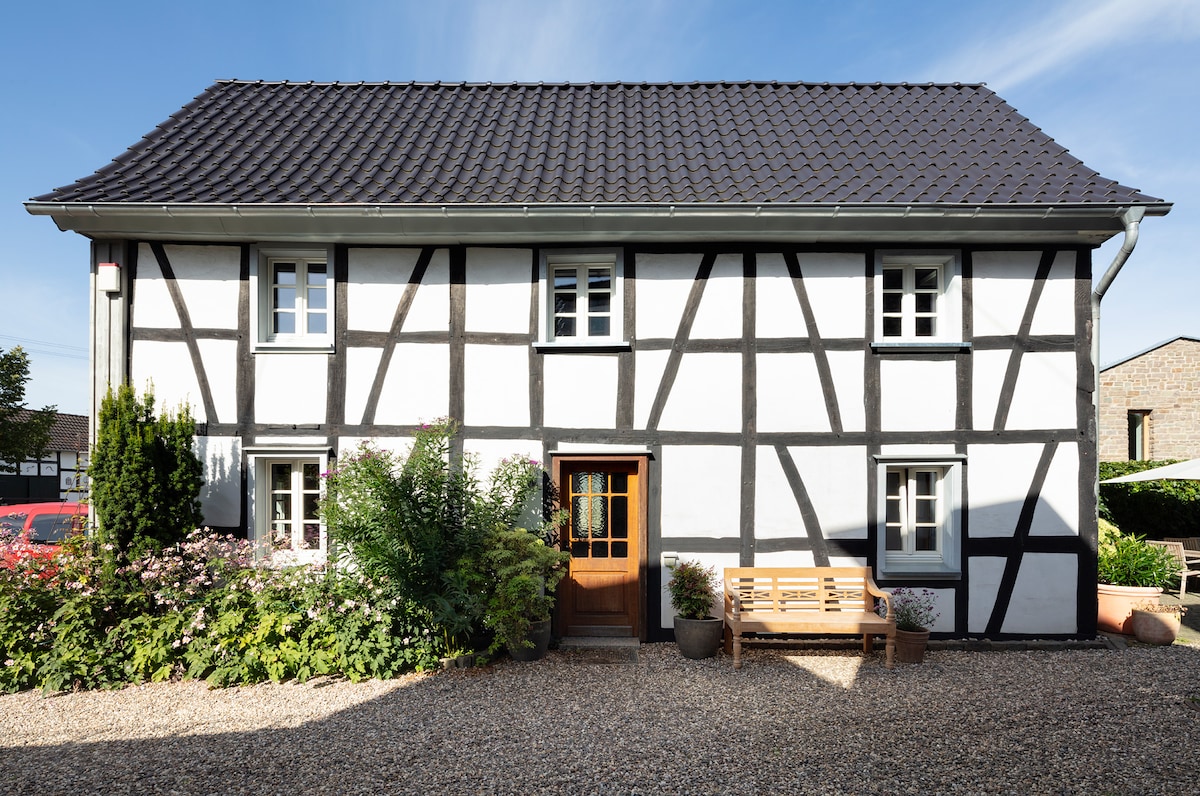 The exterior of a lovingly restored 18th-century half-timbered house is showcased, featuring a black-and-white facade with multiple windows. A gravel pathway leads to the wooden front door, accompanied by a bench and various plants that enhance the inviting entrance.