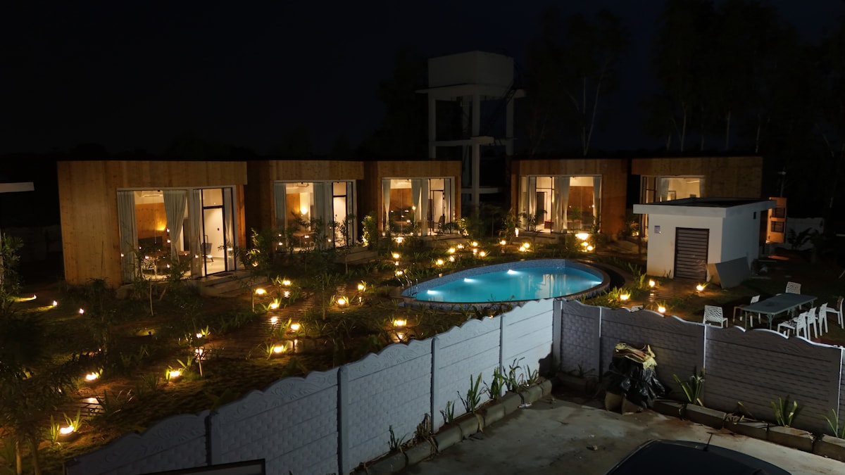 An overview of the property at night showcases five wooden houses surrounding a softly lit pool. Pathways are illuminated with warm lights, adding to the serene atmosphere. Outdoor seating is visible, enhancing the inviting environment for relaxation and social gatherings.