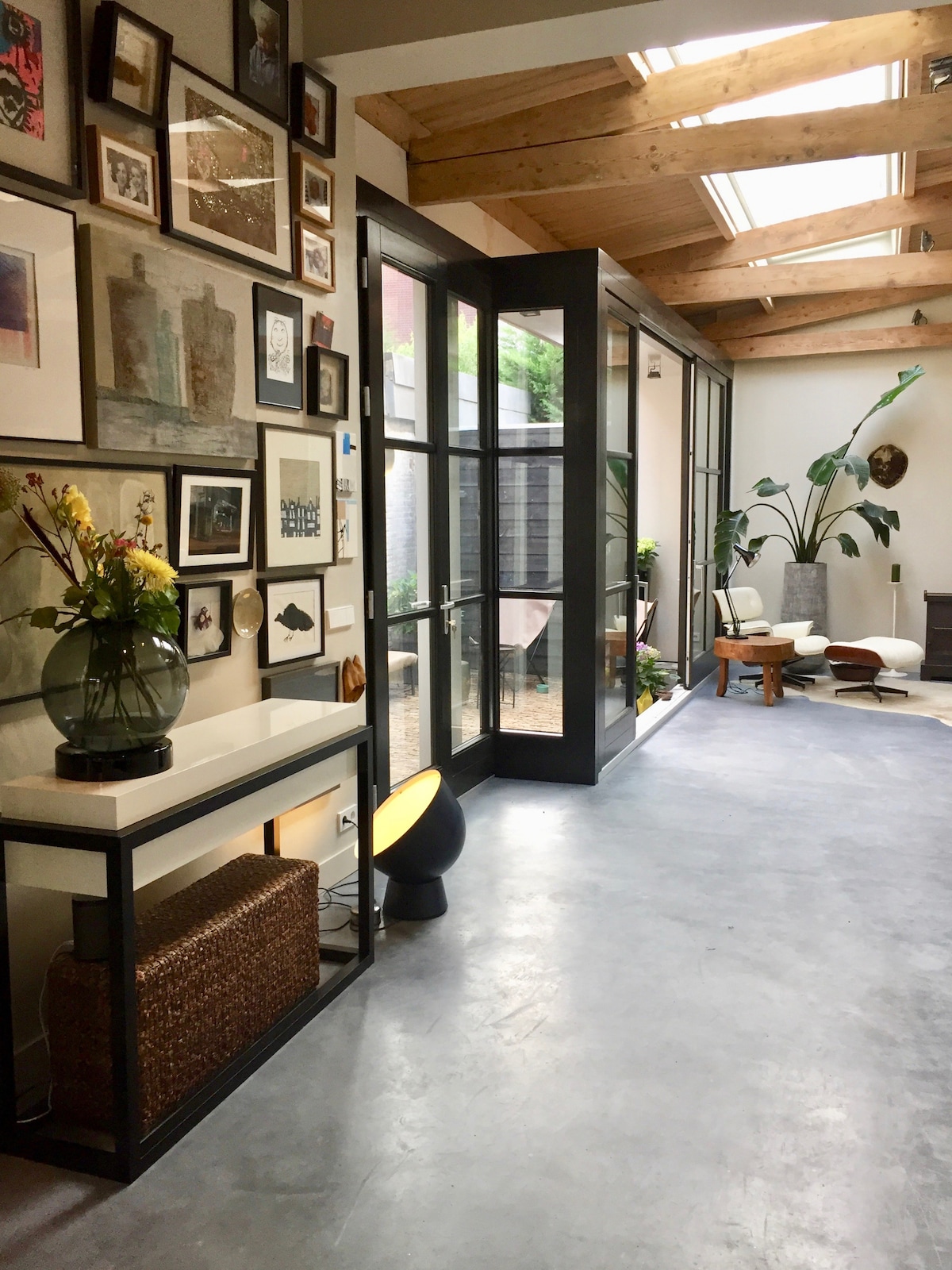 A welcoming hallway showcases a diverse gallery wall adorned with various framed artworks. Natural light streams in through large glass doors, illuminating the gray concrete floor. Potted plants add a touch of greenery, while wooden beams create a warm ceiling above.
