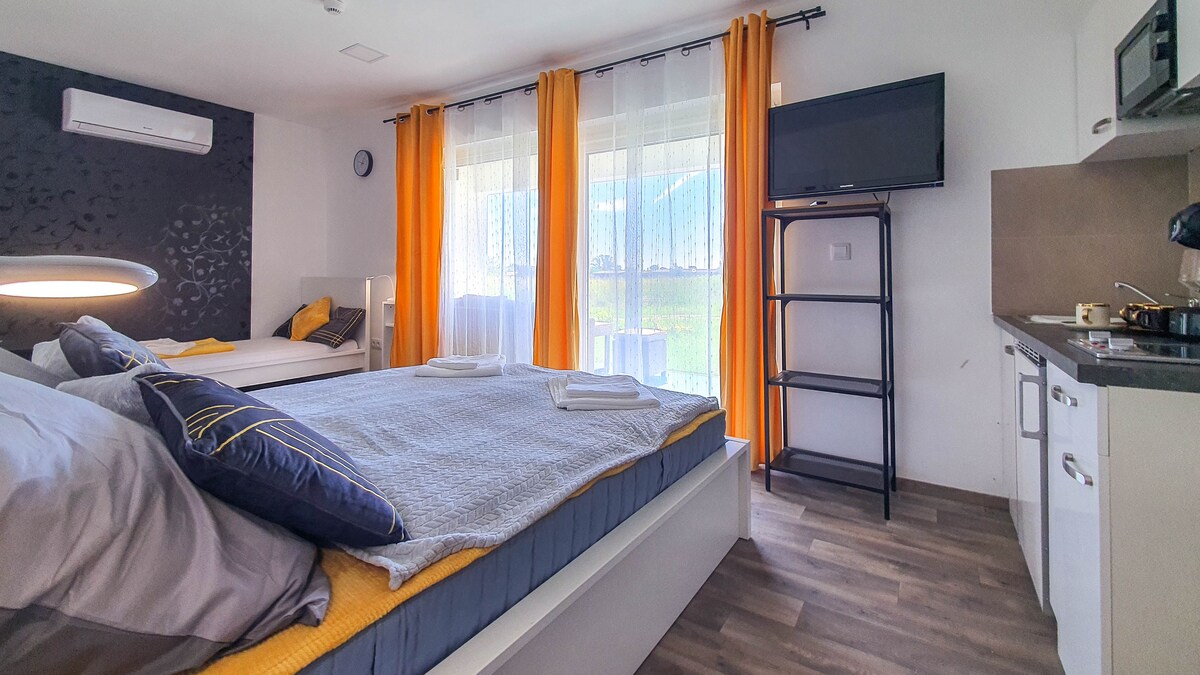 A spacious bedroom features a large bed with layered bedding and a smaller bed nearby. Natural light streams through sheer curtains, illuminating the room's modern design. A kitchenette and a small shelving unit are visible, enhancing functionality.