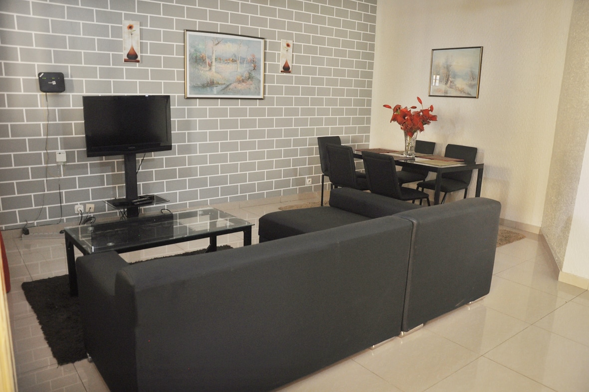 A modern living area features a dark sofa arranged in an L-shape, facing a glass coffee table. A flat-screen television is mounted on the wall, and a dining set is positioned nearby. Decorative artwork hangs on the neutral-toned walls.