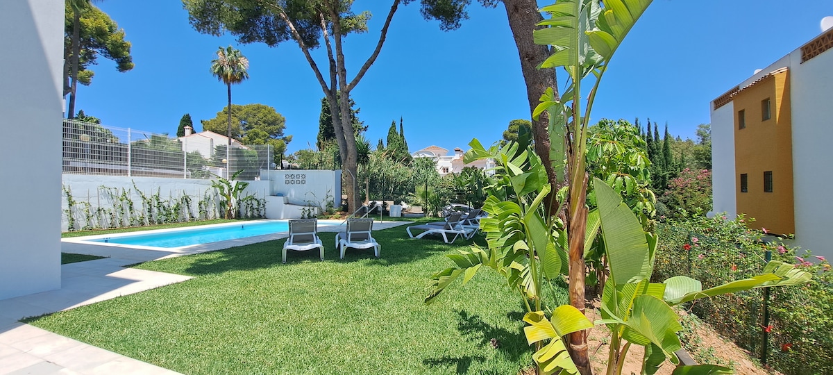 A tranquil garden area is presented with a bright green lawn, featuring lounge chairs and a clear blue pool framed by lush trees. The vibrant foliage adds a refreshing touch to the outdoor space, reflecting a serene atmosphere suitable for relaxation.