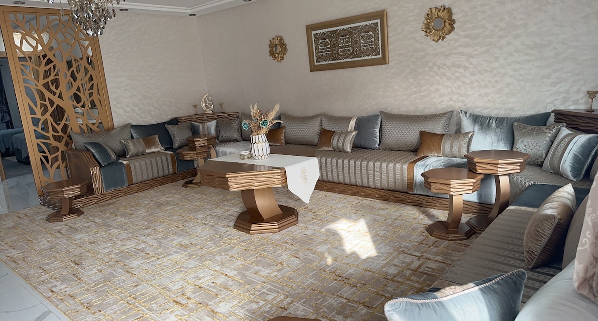 A spacious living area is presented, featuring an array of plush couches arranged around a central table. Decorative cushions in varying textures complement the neutral tones. A light-colored area rug adds warmth, while subtle wall decorations enhance the elegant ambiance.