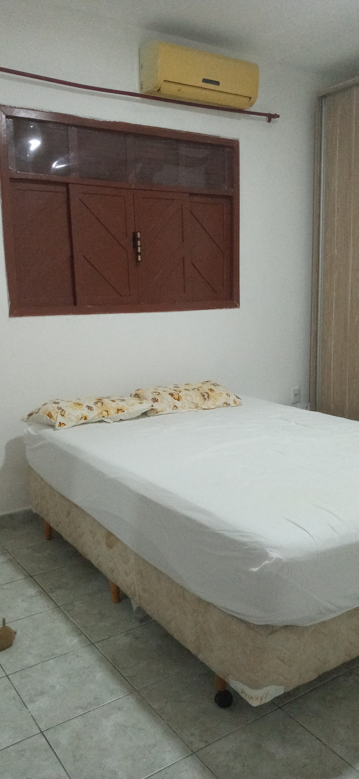 A spacious bed is set against a neutral wall, featuring light bedding and decorative pillows. A window with wooden shutters allows natural light into the room, while a beige and white patterned floor adds warmth to the space.