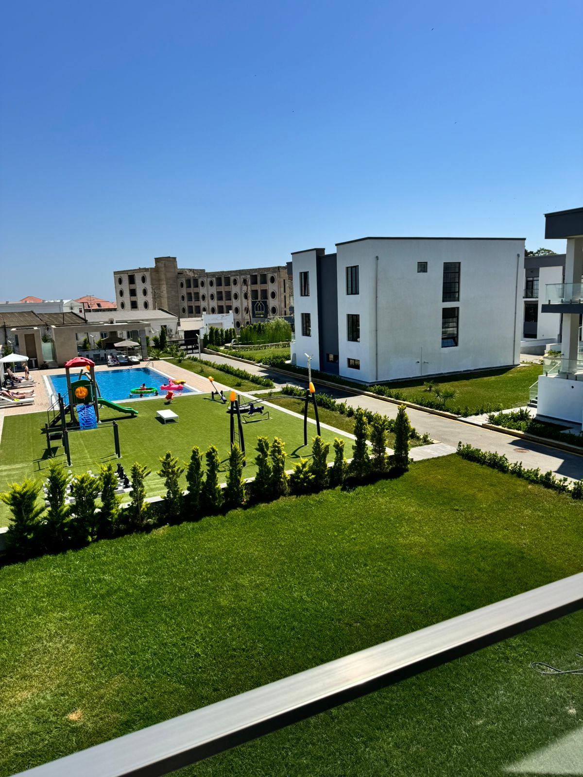 A view from the balcony showcases a large outdoor area featuring a swimming pool surrounded by lounge chairs. Colorful play structures and well-maintained grass lawns create an inviting atmosphere, while modern buildings are positioned in the background.