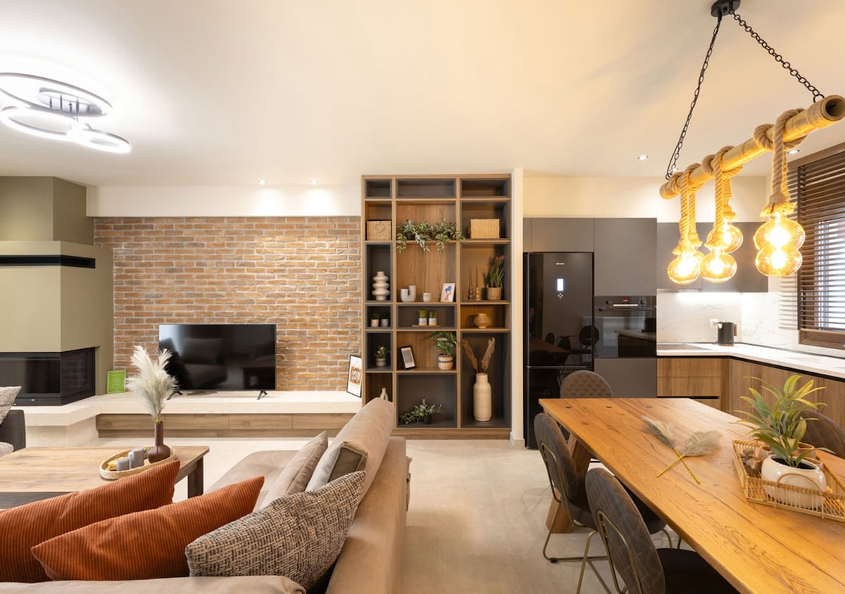 A modern living space features a cozy seating area with a light-colored sofa and decorative pillows. A rustic dining table with black chairs is illuminated by a unique light fixture. A brick wall adds texture to the room, while a dark refrigerator is visible in the kitchen area.