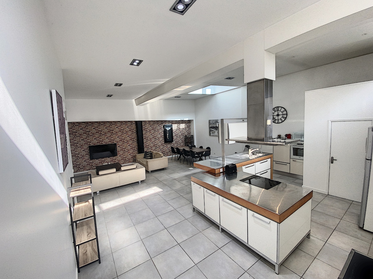 A spacious main area is highlighted by a central kitchen island with a stovetop, surrounded by modern cabinetry. A dining table for eight is positioned nearby, complemented by a comfortable living space featuring two sofas and a wall-mounted television against a decorative backdrop.