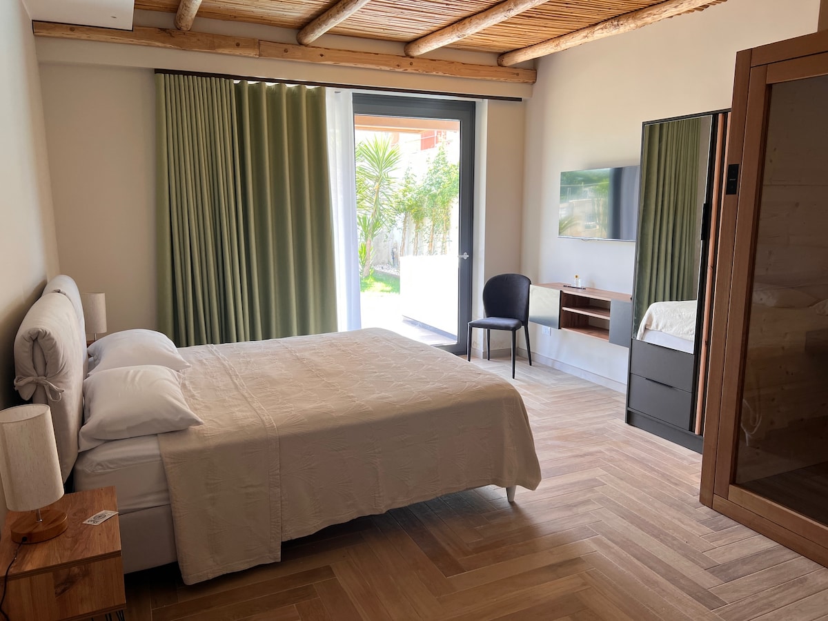 A spacious sleeping area features a large bed with neatly arranged linens. Light streams in through floor-to-ceiling doors, leading to a garden outside. A sleek TV is mounted on the wall across from a dark wooden dresser, while a simple chair is positioned nearby.