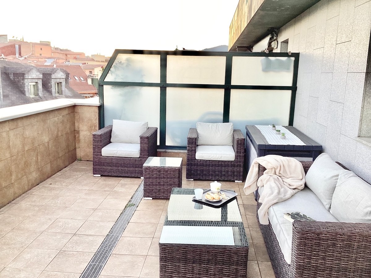 An outdoor terrace is furnished with a combination of rattan seating arrangements, including two armchairs and a spacious sofa, complemented by a glass coffee table. A tray with refreshments is placed atop the table, while the view of the surrounding rooftops can be seen in the background.