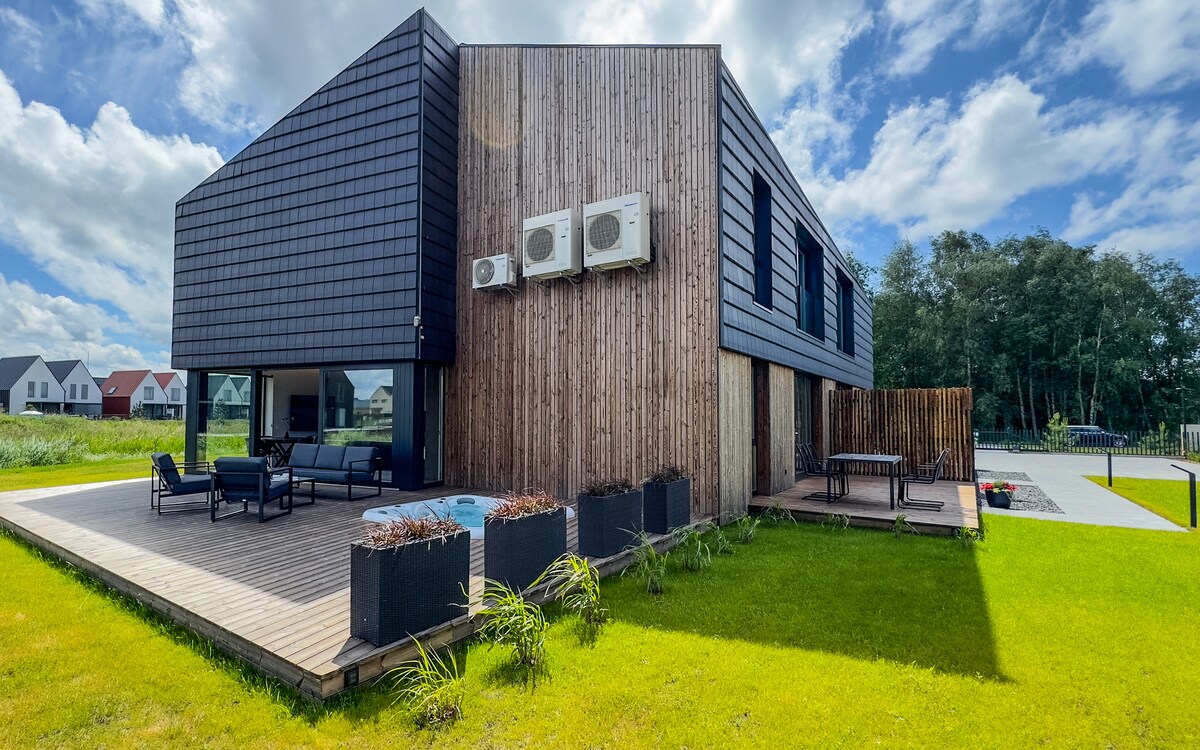 An exterior view of a two-story building featuring a contemporary design with a mix of dark siding and natural wood accents. A wooden deck extends from the building, complemented by landscaped green grass. Outdoor seating is visible, and air conditioning units are mounted on the facade.