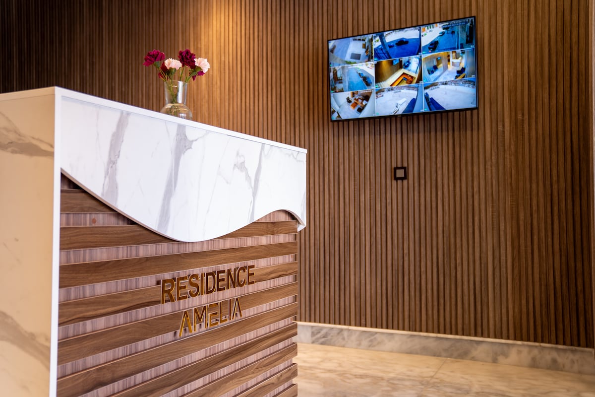 The reception area features a sleek front desk with elegant wood paneling and the name 'Residence Amelia' prominently displayed. A floral arrangement adds a touch of color, while a wall-mounted television displays security footage, ensuring a secure environment for guests.