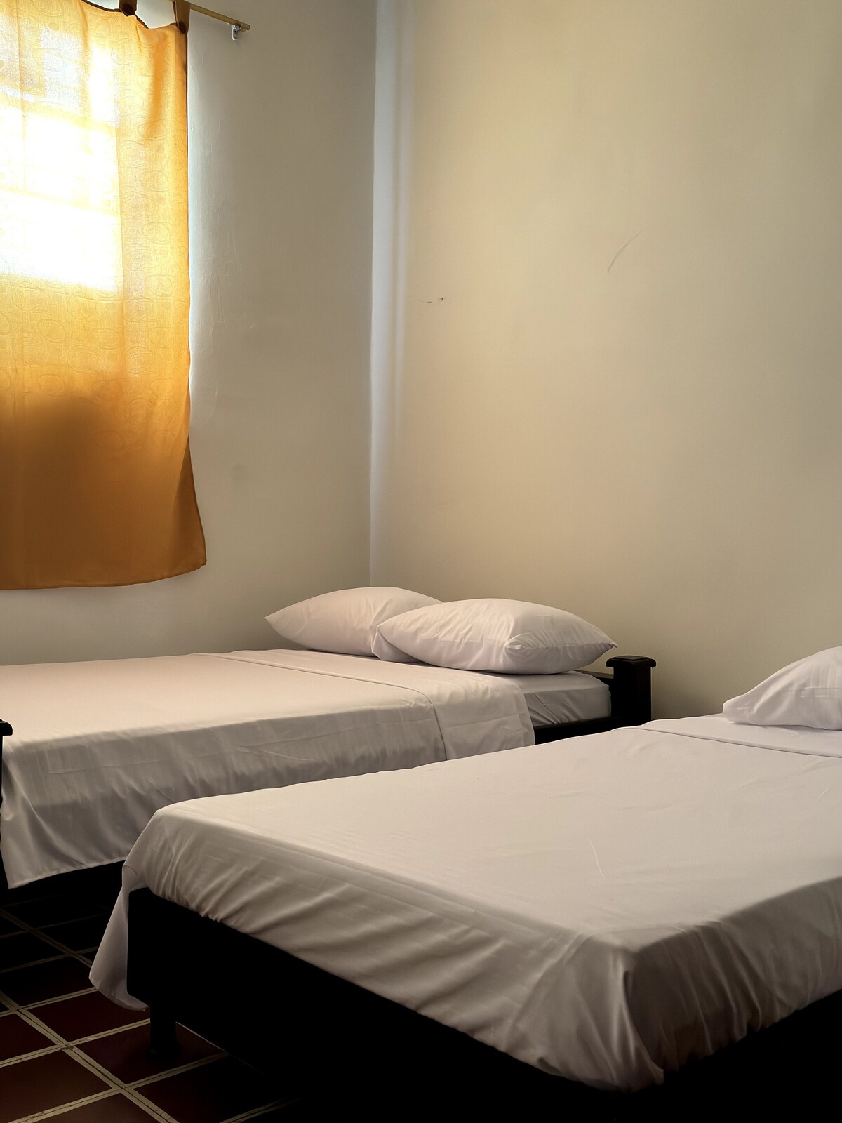 The sleeping area features two spacious beds with white linens and pillows. A window allows natural light to illuminate the room, with sheer curtains gently fluttering in the breeze. The walls are painted in soft tones, contributing to a calm environment.