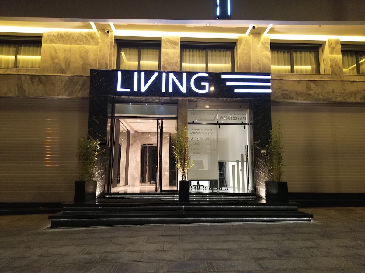 The entrance to the apartment building is showcased, featuring a sleek design with a modern sign displaying 'LIVING.' Elegant marble finishes and well-lit pathways provide an inviting approach. Glass doors lead into the interior, surrounded by subtle greenery that enhances the contemporary aesthetic.
