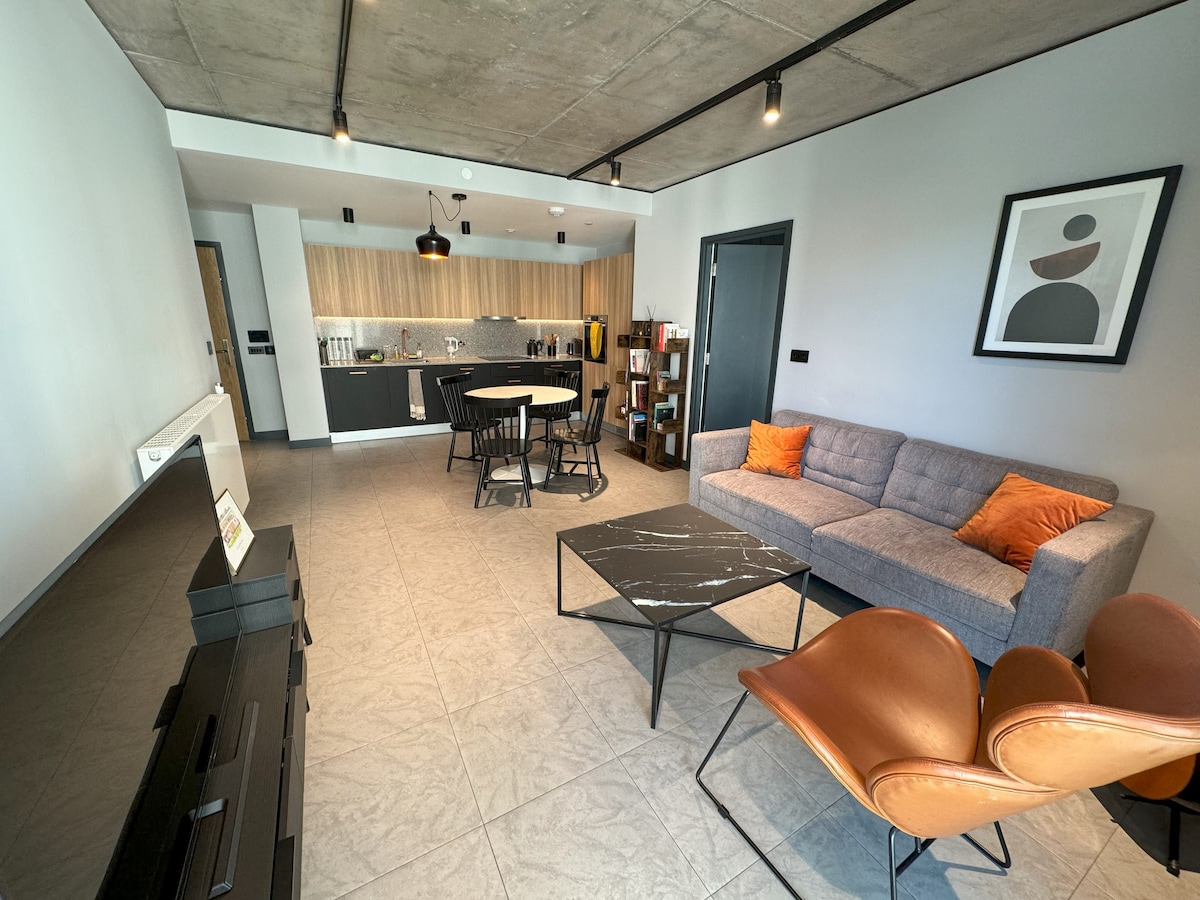 An open living area is highlighted by modern furniture, including a sectional sofa with orange cushions and stylish armchairs. A rectangular coffee table is positioned centrally, while a dining table accommodates four. The kitchen area features sleek cabinetry and is well-lit by overhead lighting.