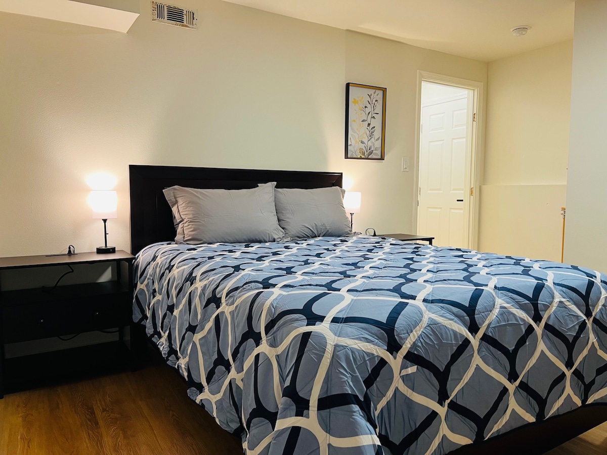 A well-appointed room showcases a queen-sized bed dressed in a patterned blue and white quilt. Nightstands with lamps flank the bed, providing soft illumination. Light wood flooring complements the neutral walls, and a framed piece of wall art adds a touch of decor. A door leads to an adjacent area.