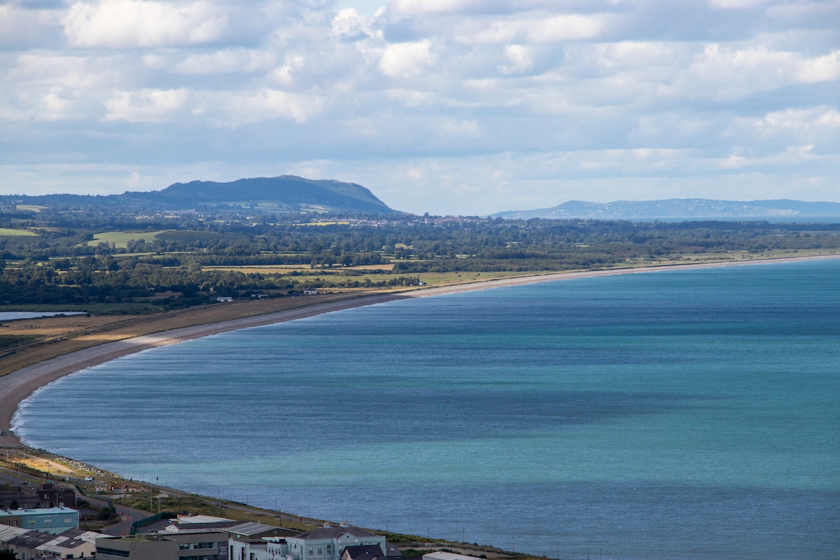 A sweeping coastal view showcases a sandy beach stretching along the shoreline, with gentle waves lapping against the shore. Rolling green hills rise in the background, framed by a partly cloudy sky, while the sea exhibits a gradient of blue shades reflecting natural light.