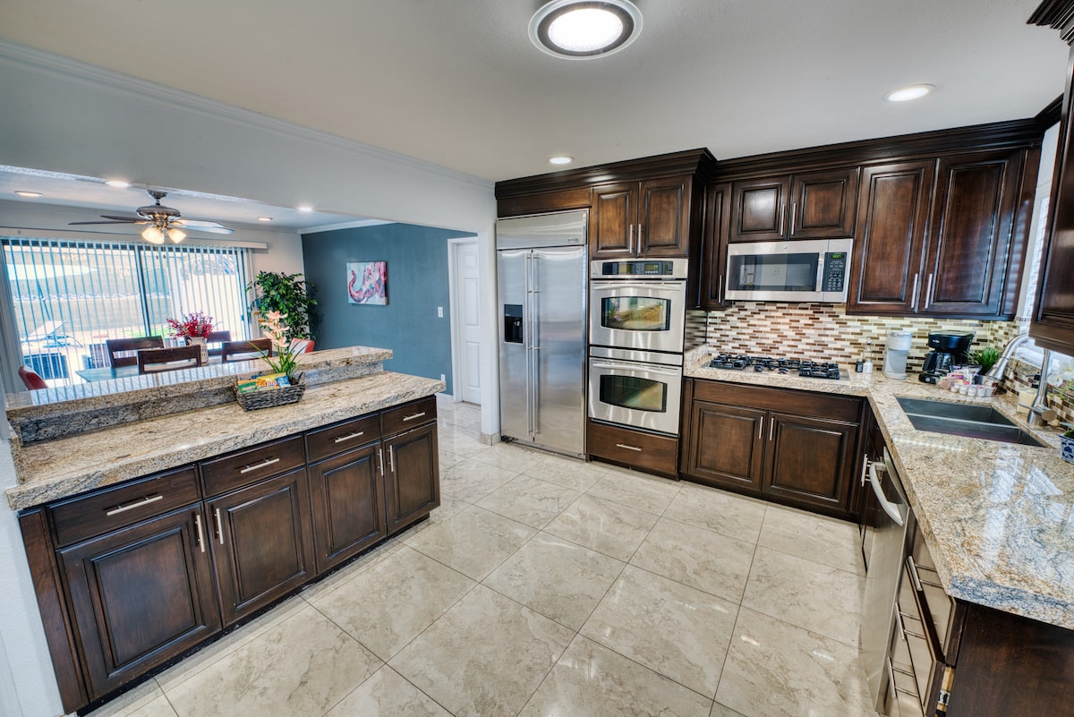 A modern kitchen features dark wooden cabinetry paired with granite countertops. Stainless steel appliances, including a refrigerator and double oven, are visible. Tile flooring reflects light from overhead fixtures and natural sunlight. An adjacent dining area is partially visible through glass sliding doors.