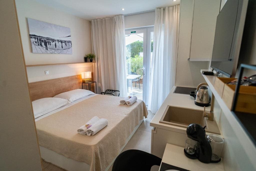 A cozy studio is depicted, featuring a spacious bed with light linens and neatly arranged towels. A kitchenette is visible, equipped with a coffee machine and a kettle. Natural light enters through a glass door that leads to a terrace, while sheer curtains provide a soft touch.