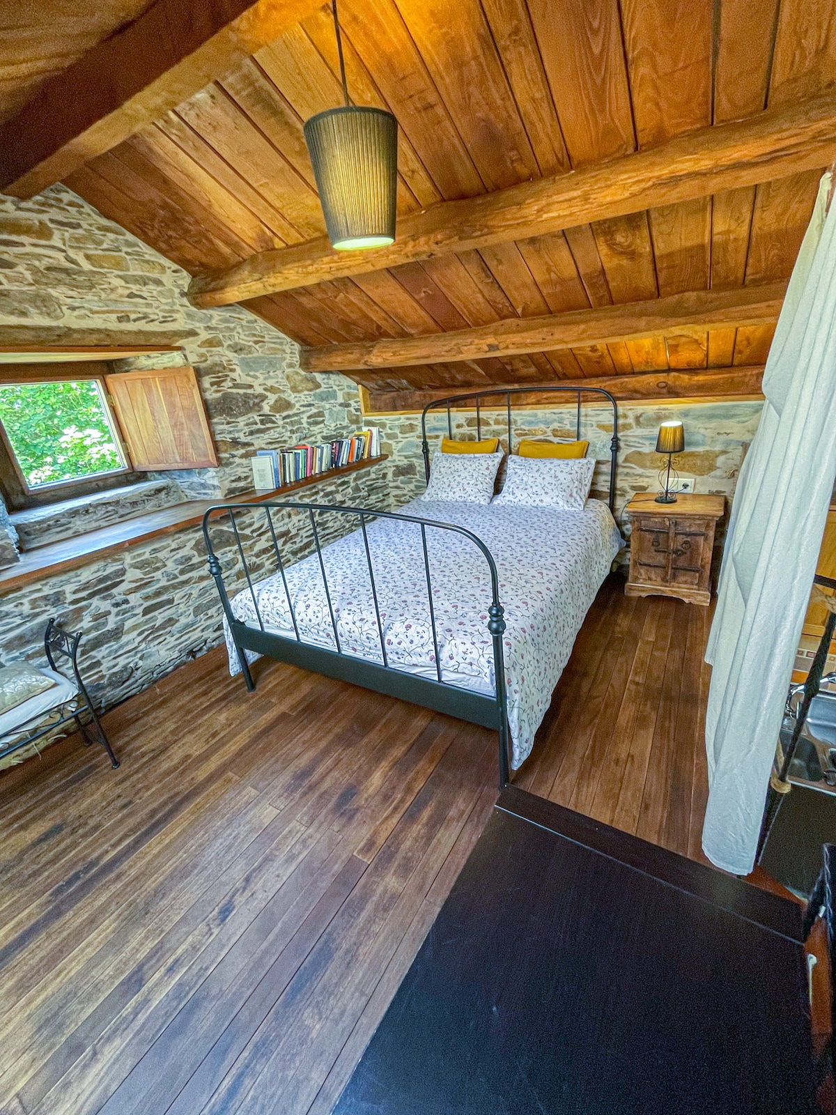 An inviting bedroom features a wrought iron bed dressed in floral bedding. Warm wood beams and stone walls contribute to the rustic charm. A small bedside table holds a lamp, while a window offers natural light and a view of the outdoors.