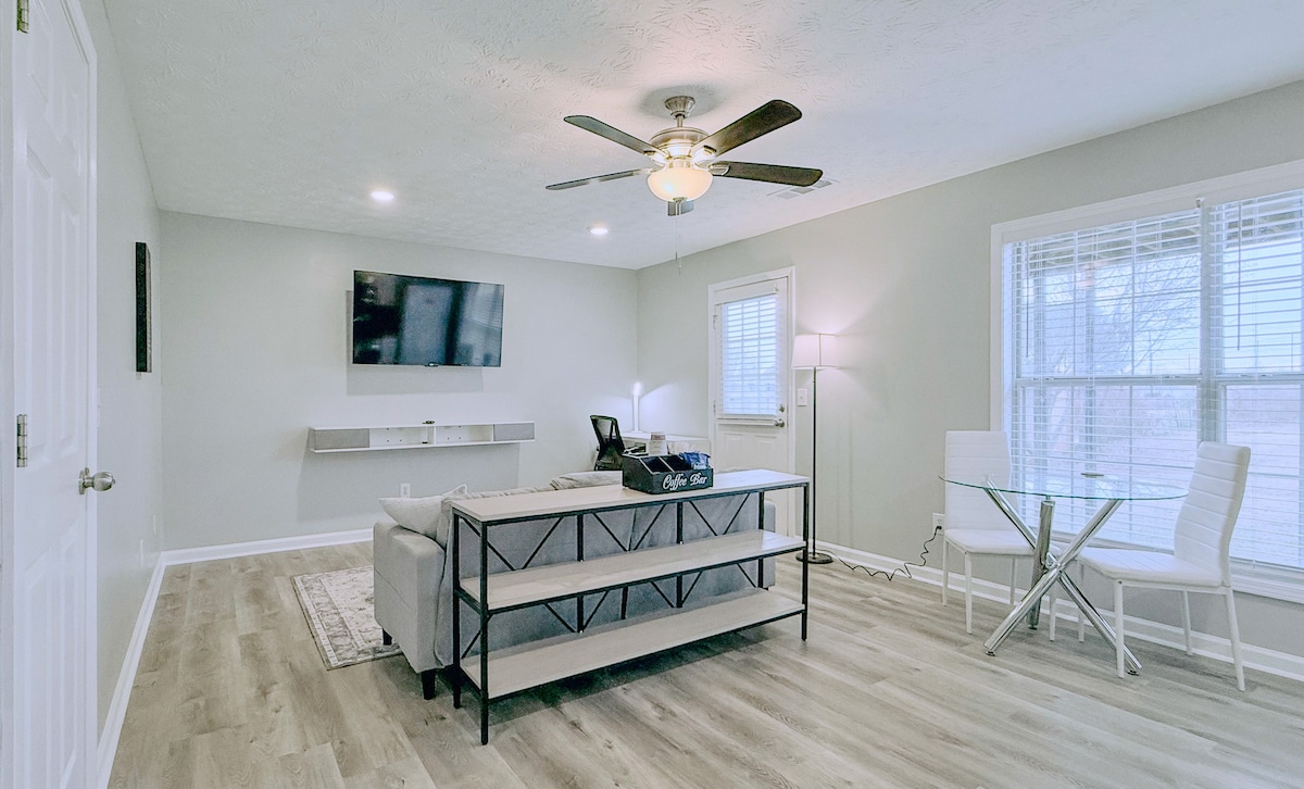 The living room features a light-colored sofa with a sleek coffee table in front, complemented by a modern ceiling fan. A wall-mounted TV is positioned above a minimalistic shelf. Natural light fills the space through large windows, while a glass dining table is set for two.