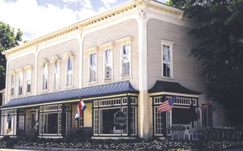 The elegant exterior of the Inn features a well-maintained facade with large windows and decorative trim. Flags adorn the entrance, while an inviting seating area is visible outside, surrounded by flower beds that enhance the welcoming ambiance of the property.