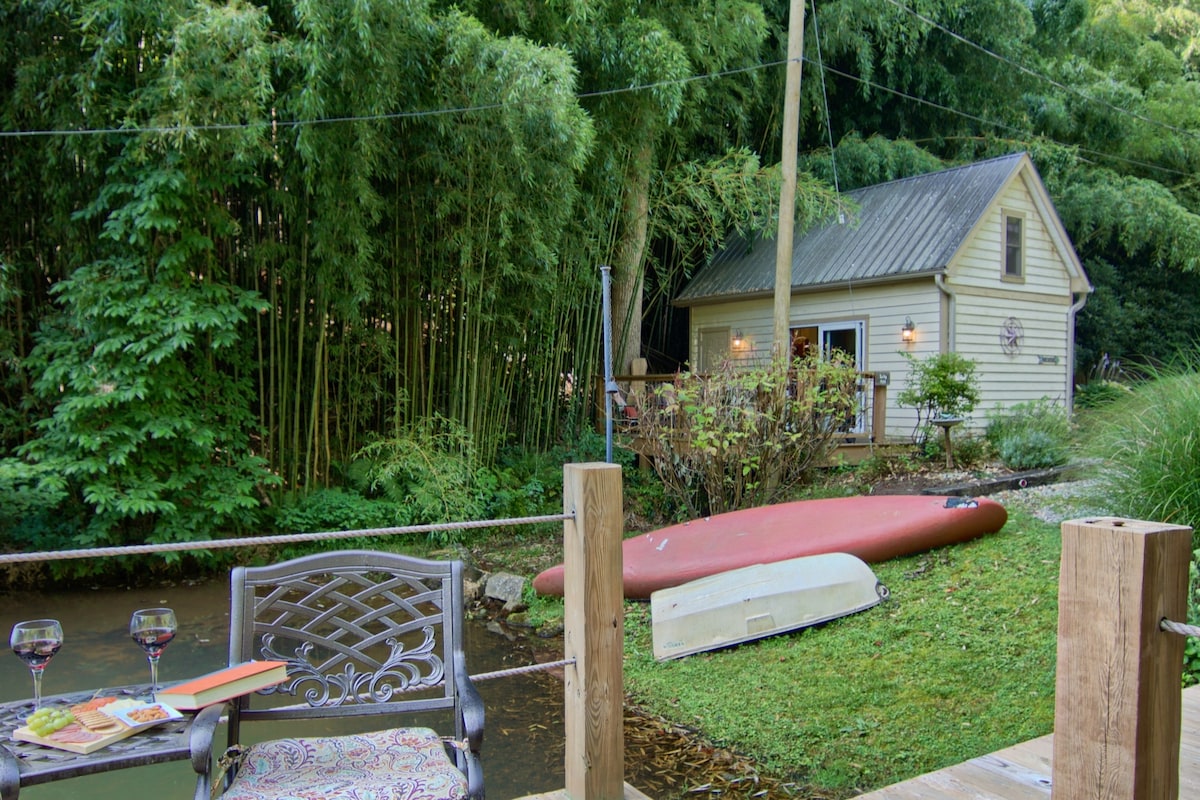A cozy cottage is nestled among greenery, with a mountain stream flowing beside it. A kayak rests on the grass near the water's edge. Two glasses of wine and a decorative plate are positioned on a wooden ledge, inviting relaxation in a natural setting.